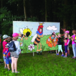 Eine Gruppe Kinder steht im Freien vor einer großen Tafel mit Comic-Bienen und Blumen und hört einer Frau zu, die teilweise als Biene verkleidet ist.