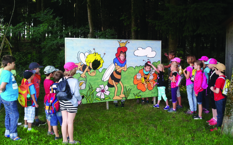 Eine Gruppe Kinder steht im Freien vor einer großen Tafel mit Comic-Bienen und Blumen und hört einer Frau zu, die teilweise als Biene verkleidet ist.