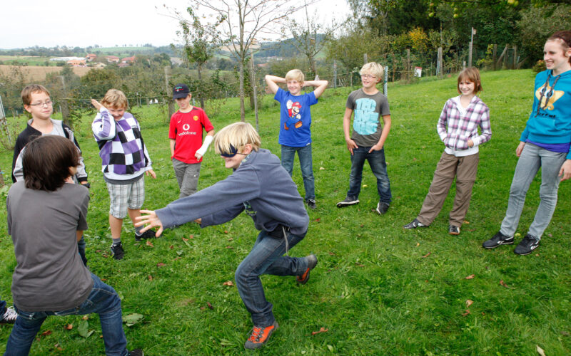 Eine Gruppe Kinder steht an einem bewölkten Tag in einem Kreis auf dem Rasen und beobachtet zwei Kinder, die in der Mitte ein Spiel spielen.