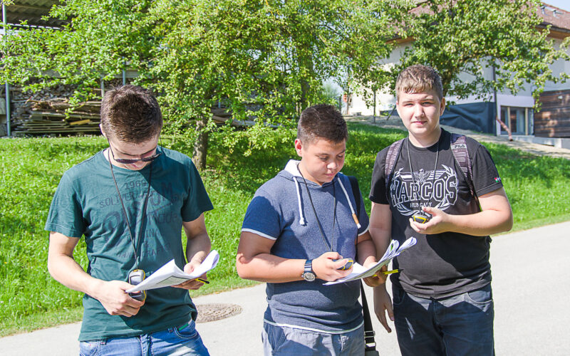 Drei junge Männer stehen an einem sonnigen Tag im Freien, schauen auf Papiere und halten Stifte in der Hand, im Hintergrund sind Bäume und Gebäude zu sehen.