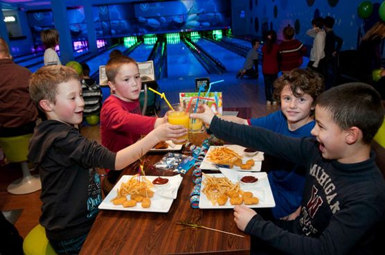 Vier Jungen sitzen an einem Tisch in einer Bowlingbahn, stoßen mit Getränken an und essen Pommes und Chicken Nuggets.