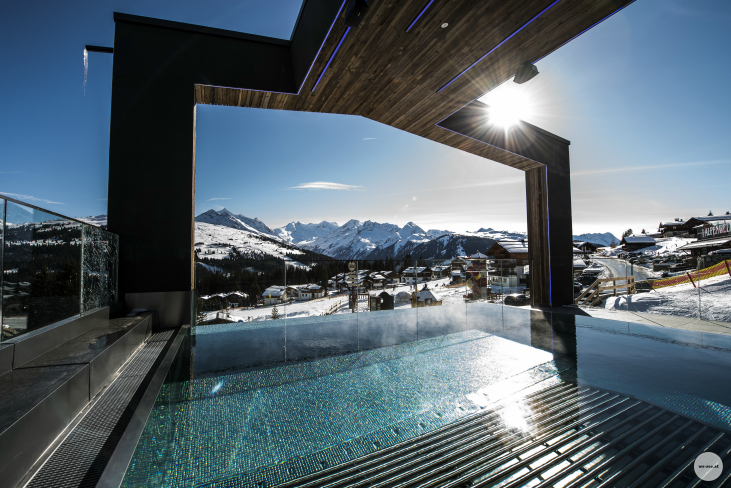 Infinity-Pool im Freien mit Blick auf die Berge, moderne architektonische Struktur über dem Wasser, strahlender Sonnenschein, schneebedeckte Landschaft im Hintergrund.