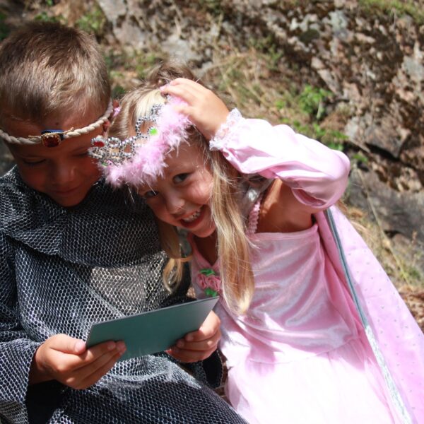 Ein Junge und ein Mädchen in Kostümen, der eine als Ritter, der eine Karte liest, und die andere als Prinzessin, die lächelt und ihre Krone zurechtrückt, sitzen im Freien zusammen.