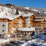 Ein großes Alpenhotel mit Holzbalkonen ist umgeben von Schnee, parkenden Autos und verschneiten Bergen unter einem strahlend blauen Himmel.