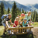 Eine fünfköpfige Familie, darunter drei Kinder, sitzt auf einem Holzkarren in einer grasbewachsenen Berglandschaft mit Bäumen und Hügeln im Hintergrund.