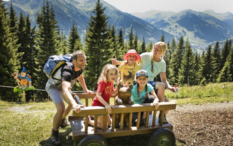Eine fünfköpfige Familie, darunter drei Kinder, sitzt auf einem Holzkarren in einer grasbewachsenen Berglandschaft mit Bäumen und Hügeln im Hintergrund.