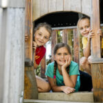 Drei Kinder im Inneren eines hölzernen Spielgerüsts, die lächelnd durch eine offene Tür hinausschauen.