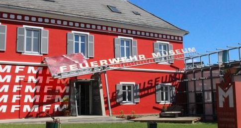Ein rotes Gebäude mit grauen Fensterläden beherbergt das Feuerwehrmuseum; ein großes Schild an einer Leiter zeigt den Namen des Museums über dem Eingang an.