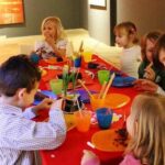 Eine Gruppe von Kindern sitzt um einen Tisch mit bunten Tellern und Tassen, isst und unterhält sich in einem hell erleuchteten Raum.