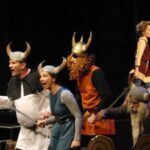 Sechs Personen in Wikingerkostümen, einige mit Hörnerhelmen, treten während eines Theaterstücks lebhaft auf.