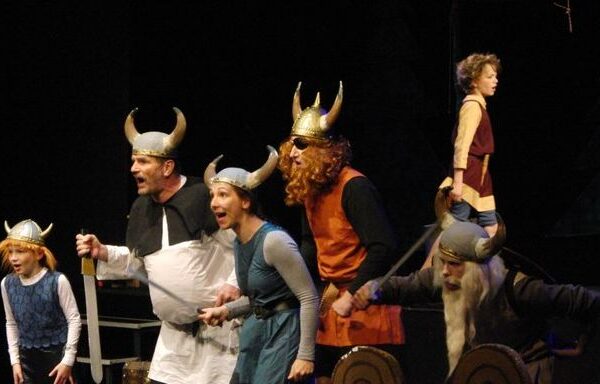 Sechs Personen in Wikingerkostümen, einige mit Hörnerhelmen, treten während eines Theaterstücks lebhaft auf.