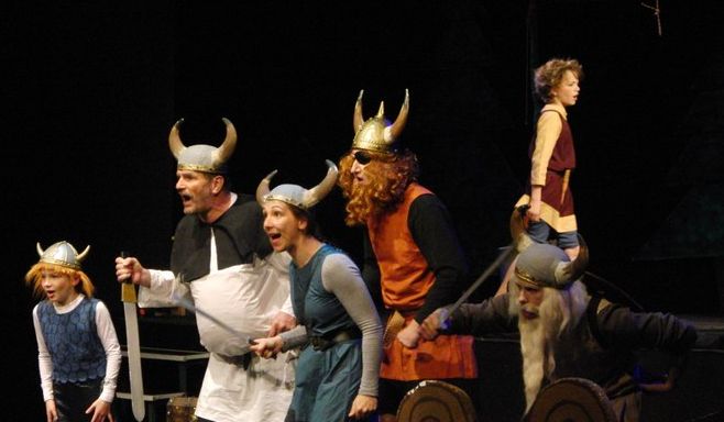 Sechs Personen in Wikingerkostümen, einige mit Hörnerhelmen, treten während eines Theaterstücks lebhaft auf.