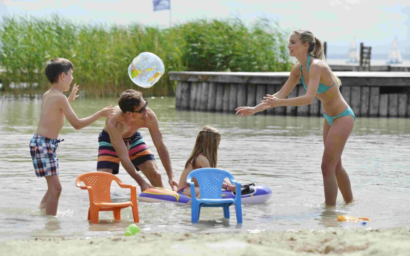 Eine Familie mit zwei Kindern spielt an einem sonnigen Tag mit aufblasbarem Spielzeug und Plastikstühlen im flachen Wasser eines Strandes.