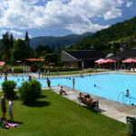 Außenpool mit schwimmenden und entspannenden Menschen; Rasenfläche mit Sonnenschirmen und Liegestühlen; Gebäude und grüne Hügel im Hintergrund unter einem teilweise bewölkten Himmel.