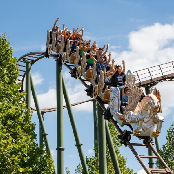 Eine Gruppe von Menschen mit erhobenen Armen fährt auf einer Achterbahn in Form von Pferden eine Strecke im Freien hinunter, umgeben von grünen Bäumen und einem blauen Himmel.