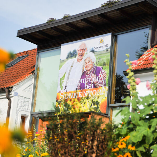 Ein Gebäude mit rotem Ziegeldach trägt ein großes Schild mit einem älteren Ehepaar und der Aufschrift "Willkommen bei Sonnentor", umgeben von Blumen und Grünpflanzen.