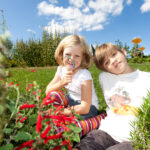 Zwei Kinder sitzen an einem sonnigen Tag im Gras eines Gartens; eines hält sich eine Lupe vors Gesicht, beide sind von roten Blumen und Grün umgeben.