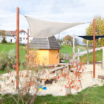 Ein kleiner Spielplatz mit einem Sandkasten, Holzkonstruktionen und einer gelben Hütte, umgeben von Gras, Bäumen und Häusern im Hintergrund.