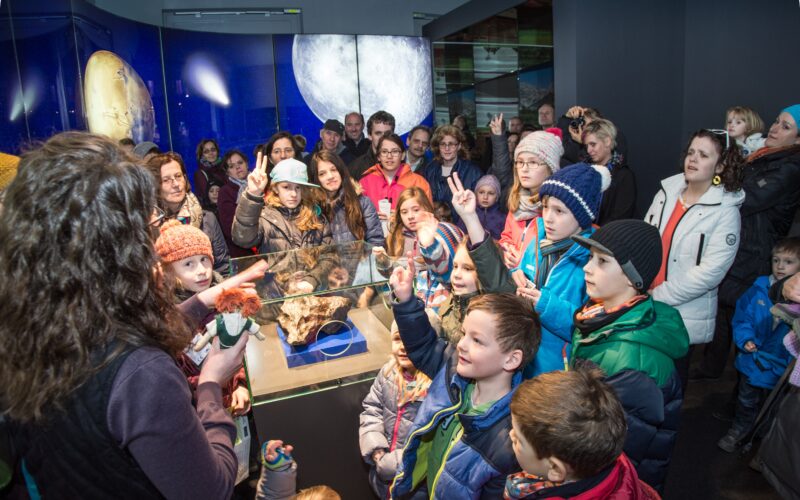 Eine Gruppe von Kindern und Erwachsenen versammelt sich um eine Vitrine in einer Museumsausstellung, während ein Führer ein Objekt zeigt und zur Menge spricht.