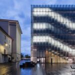 Ein modernes Glasgebäude mit beleuchteten horizontalen Linien steht zwischen älteren Gebäuden in einem feuchten, leeren Innenhof in der Abenddämmerung.