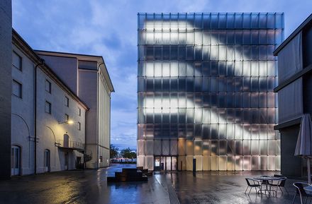 Ein modernes Glasgebäude mit beleuchteten horizontalen Linien steht zwischen älteren Gebäuden in einem feuchten, leeren Innenhof in der Abenddämmerung.