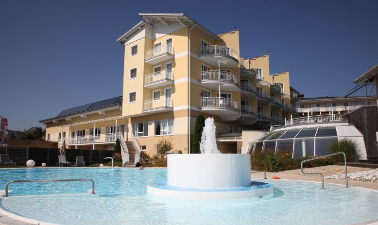 Ein modernes, gelbes Hotelgebäude mit mehreren Balkonen überblickt einen Außenpool mit einem zentralen Springbrunnen unter einem klaren blauen Himmel.