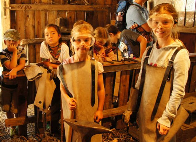 Kinder mit Schutzbrillen und Schürzen stehen in einer Werkstatt mit Schmiedewerkzeugen, einige lächeln, in einem scheunenähnlichen Holzgebäude.