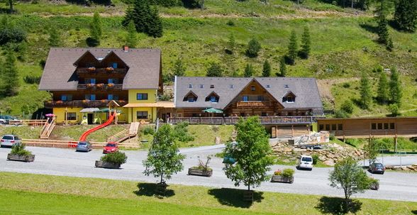 Zwei große Holzhütten mit Balkonen und einer roten Spielplatzrutsche stehen auf einem grasbewachsenen Hügel, mit mehreren geparkten Autos und Bäumen im Vordergrund.