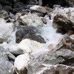 Ein kleiner Wasserlauf fließt über und zwischen großen, unregelmäßigen Felsen und Blöcken, die von einigen Baumzweigen umgeben sind.