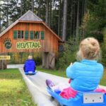 Zwei Kinder fahren auf blauen Schlitten eine Betonrutsche hinunter zu einer Holzhütte mit der Aufschrift "HEUSTADL" in einem Waldgebiet. Links ist ein gelbes Warnschild "GEFAHR!" zu sehen.