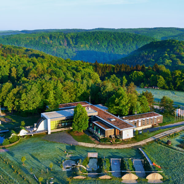 Luftaufnahme eines modernen Gebäudes, umgeben von Bäumen und Hügeln, mit Gärten, Wegen und einem Parkplatz im Vordergrund.