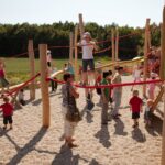 Kinder und Erwachsene treffen sich auf einem Außenspielplatz mit Holzkonstruktionen und roten Seilen, umgeben von Bäumen und einer Wiese.
