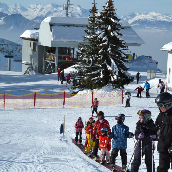 Kinder und Erwachsene in Skiausrüstung stehen in einem verschneiten Skigebiet auf einem Förderband in einer Reihe, im Hintergrund sind Berge, Bäume und Gebäude zu sehen.