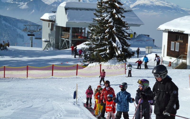 Kinder und Erwachsene in Skiausrüstung stehen in einem verschneiten Skigebiet auf einem Förderband in einer Reihe, im Hintergrund sind Berge, Bäume und Gebäude zu sehen.