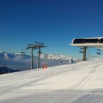 Eine schneebedeckte Skipiste mit einer Skiliftstation, Skiliftsesseln und Bergketten im Hintergrund unter einem klaren blauen Himmel.