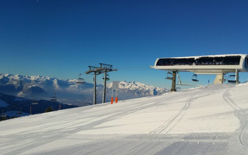 Eine schneebedeckte Skipiste mit einer Skiliftstation, Skiliftsesseln und Bergketten im Hintergrund unter einem klaren blauen Himmel.