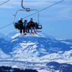 Drei Personen in Skiausrüstung fahren mit einem Sessellift über eine verschneite Berglandschaft mit klarem blauen Himmel im Hintergrund.