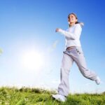 Eine Frau in weißer Kleidung geht zügig auf einer Wiese unter einem strahlend blauen Himmel mit Sonnenschein.