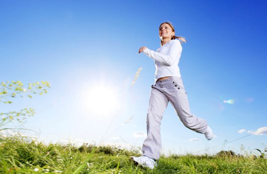 Eine Frau in weißer Kleidung geht zügig auf einer Wiese unter einem strahlend blauen Himmel mit Sonnenschein.