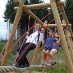 Drei Personen balancieren und gehen auf einer Hängeseilbrücke, die von Holzbalken und roten Seilen getragen wird, in einer Außenumgebung mit Bäumen im Hintergrund.