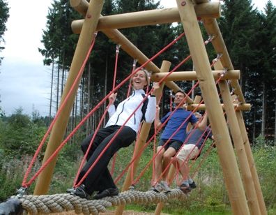 Drei Personen balancieren und gehen auf einer Hängeseilbrücke, die von Holzbalken und roten Seilen getragen wird, in einer Außenumgebung mit Bäumen im Hintergrund.