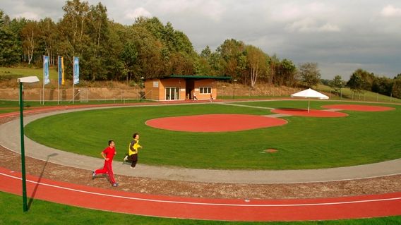 Zwei Personen joggen auf einer Laufbahn im Freien neben einem grasbewachsenen Feld mit roten Sportflächen, Bäumen und einem kleinen Gebäude im Hintergrund.