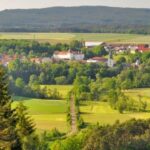 Ein kleines Dorf ist umgeben von grünen Feldern, Bäumen und bewaldeten Hügeln unter einem klaren Himmel. Eine schmale Straße führt auf das Dorf zu.