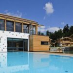 Modernes Gebäude mit großen Fenstern und Holzakzenten neben einem Außenpool, mit Bäumen und Häusern im Chalet-Stil im Hintergrund unter einem teilweise bewölkten Himmel.