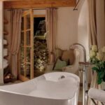 Eine moderne weiße Badewanne steht im Mittelpunkt eines gemütlichen Badezimmers mit Holzakzenten, einem Sessel, einem Strauß weißer Rosen und Glastüren nach draußen.