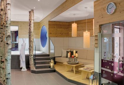 Moderne Spa-Lounge mit Holzakzenten, Birkendekor, einem Sitzbereich mit Kerzen, einer Wanduhr und einem Whirlpool, der teilweise im Hintergrund zu sehen ist.