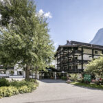 Ein mehrstöckiges Alpenhotel mit dunklen Holzbalkonen und weißen Wänden liegt zwischen Bäumen und Bergen unter blauem Himmel.