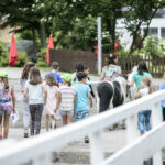 Eine Gruppe von Kindern geht gemeinsam auf einem Pfad in der Nähe eines Ponys auf einem Bauernhof spazieren, beaufsichtigt von Erwachsenen, mit Bäumen und einem weißen Zaun in der Nähe.