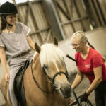 Ein junges Mädchen, das einen Helm trägt, sitzt auf einem Pferd, während ein Reitlehrer in einem roten Hemd in einer Reithalle daneben steht.