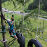 Eine Person mit Sicherheitsausrüstung navigiert durch einen Hochseilgarten, hält sich an Seilen fest und tritt auf aufgehängte Reifen, während im Hintergrund eine Graslandschaft und Bäume zu sehen sind.